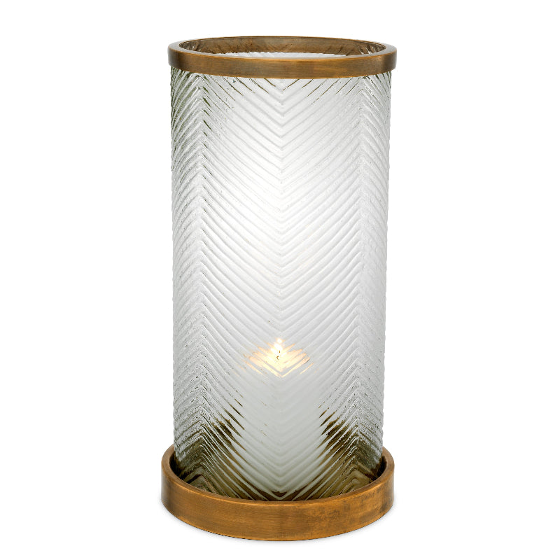 A cylindrical glass hurricane with ribbed texture and antique brass base and rim, containing a lit candle.